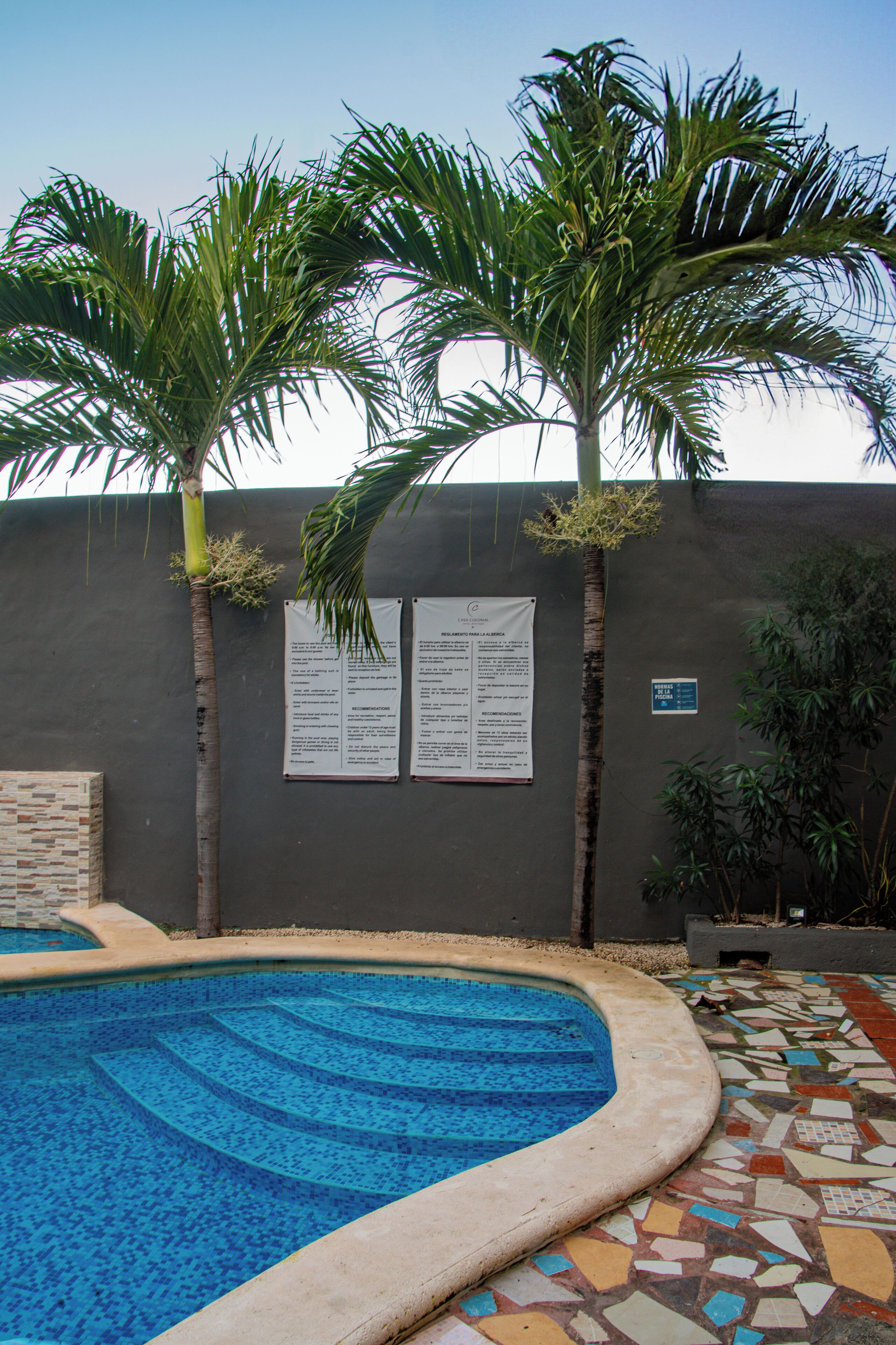 Ein Pool mit Fliesenrand, umgeben von Palmen und einer Mauer mit Hinweisschildern.