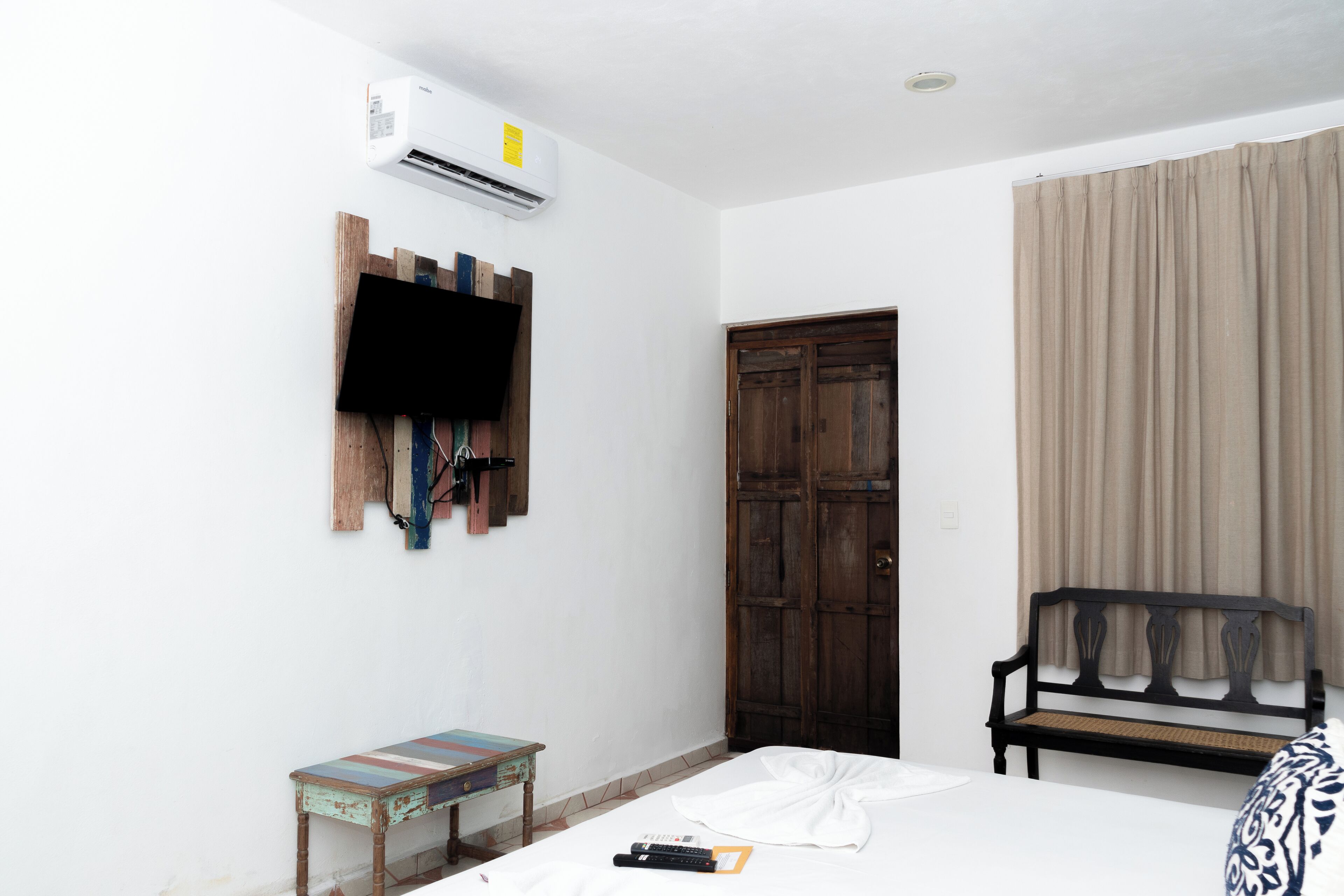 Une pièce comprenant un lit, une armoire en bois, un banc, une petite table et un téléviseur fixé au mur.