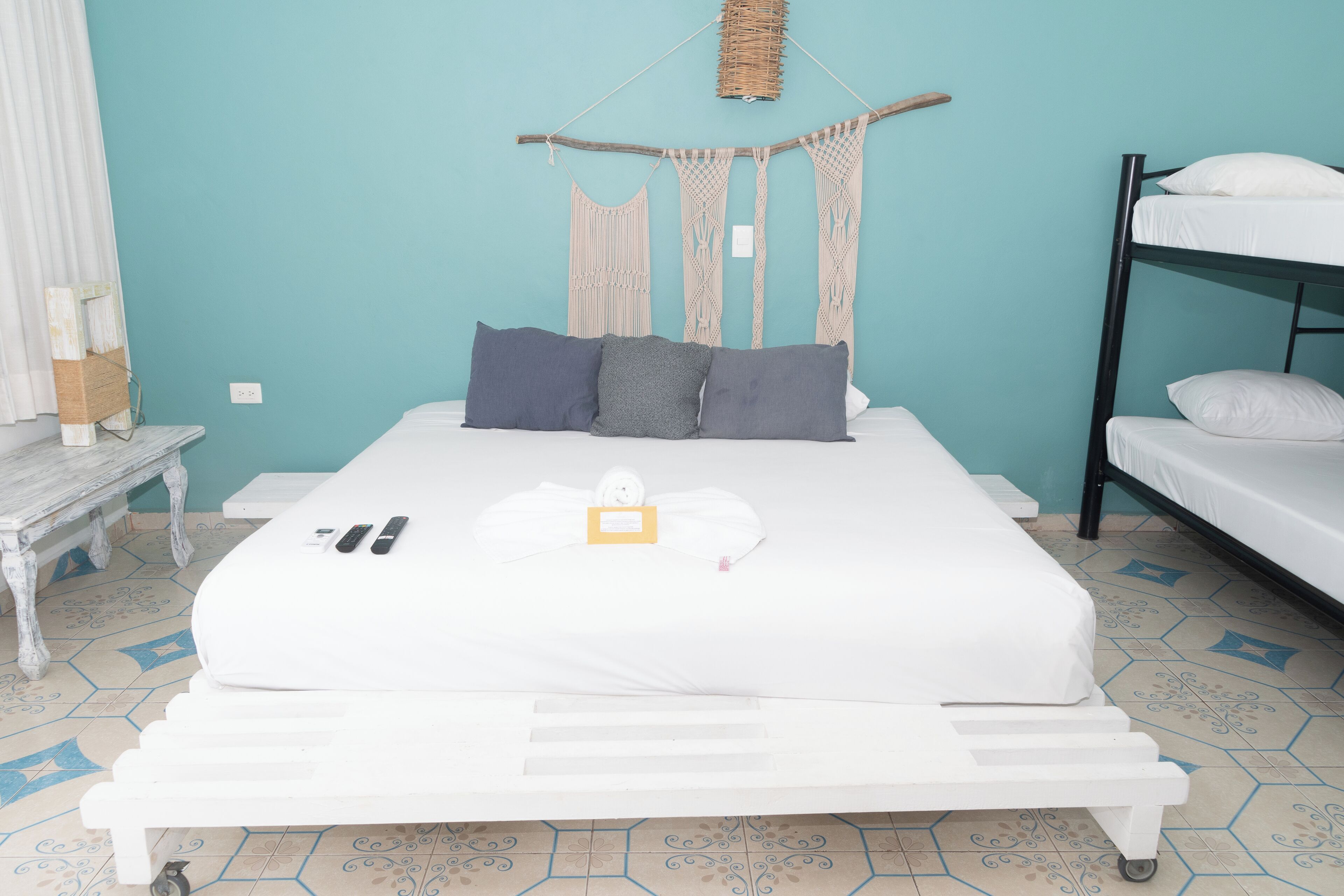 A bed with a white mattress, grey and white pillows, a bedside table with a lamp, and a bunk bed to the right.