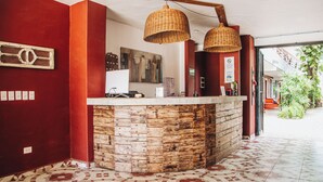 Un área de recepción con mostrador de madera, lámparas colgantes de mimbre, un cuadro enmarcado y suelo de baldosas.