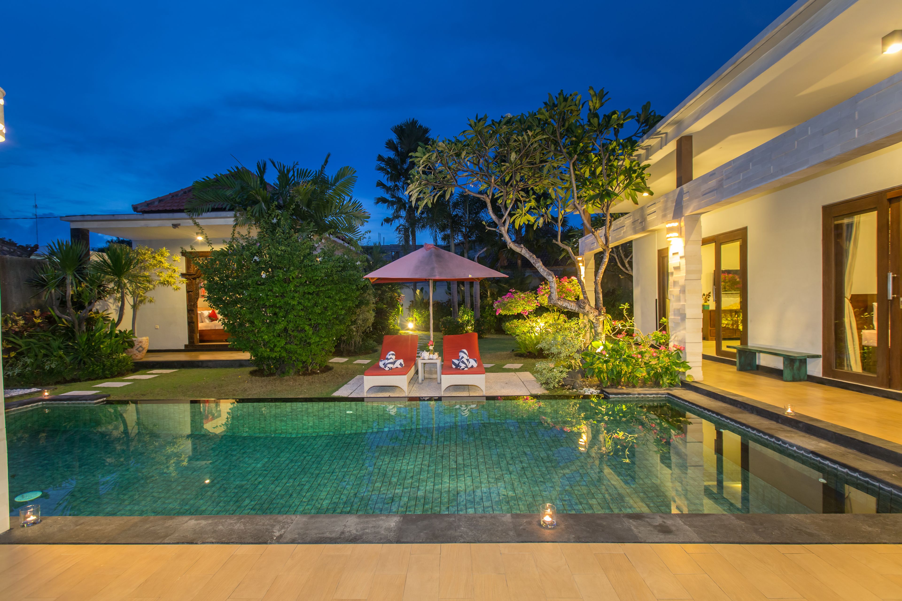 A swimming pool with lounge chairs and a small table, surrounded by greenery and a building with large windows.