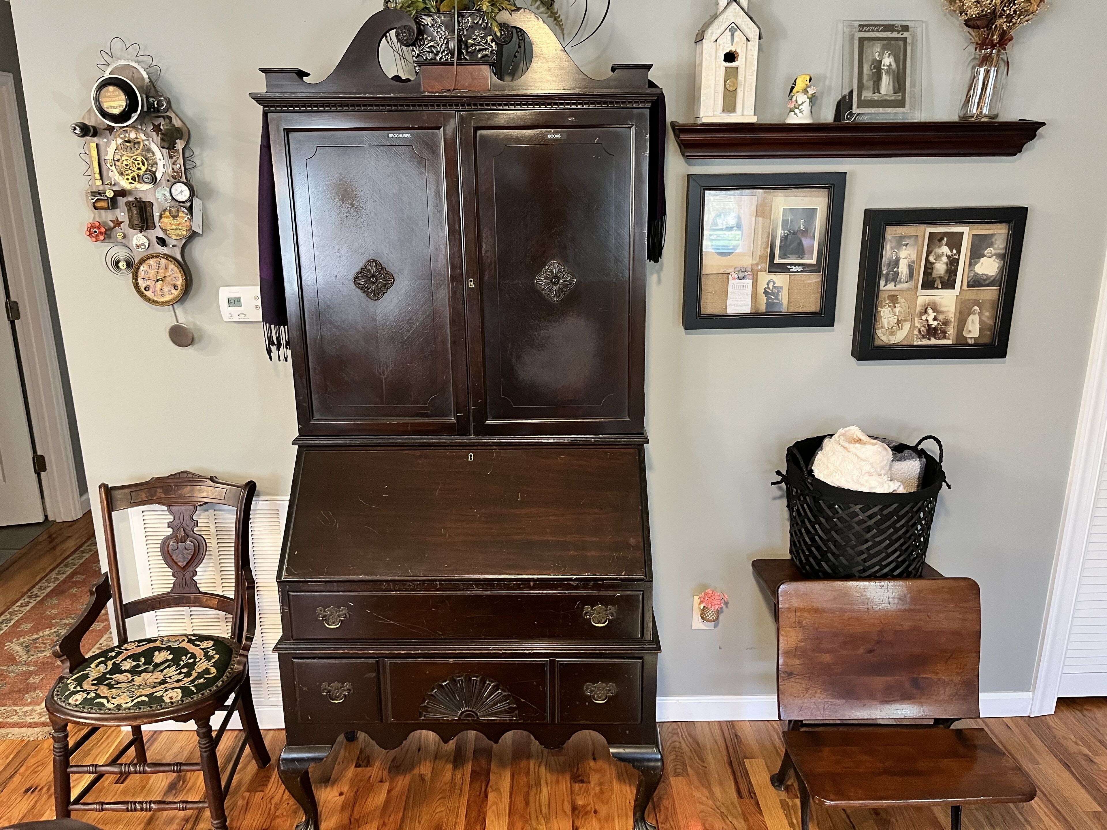 A room with a large dark wooden cabinet, a vintage chair, a small wooden table, and a wall adorned with framed pictures and a clock.