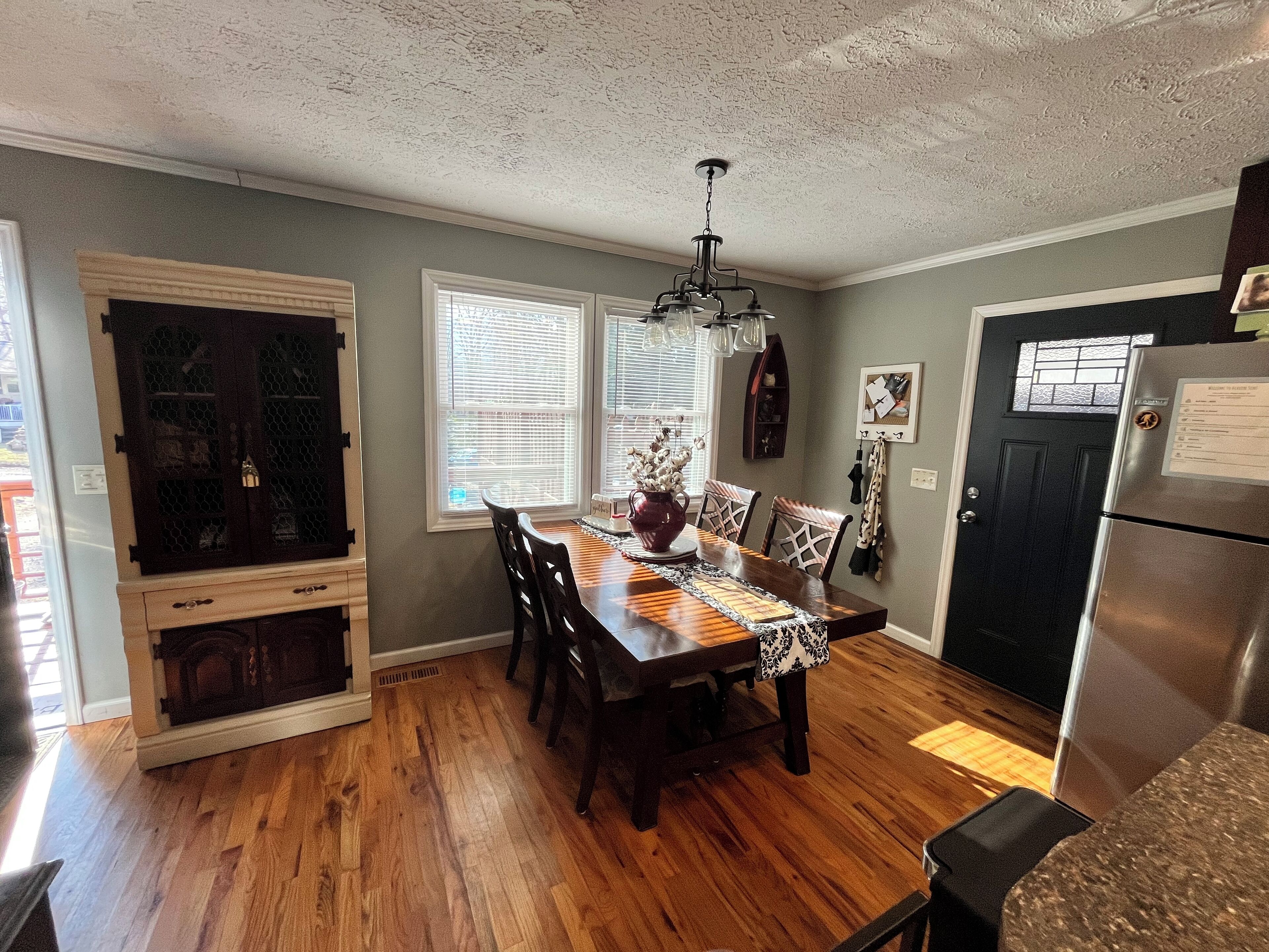 A dining area with a wooden table, chairs, and a chandelier. There is a black refrigerator and a wooden cabinet with glass doors.
