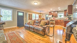 A spacious living room with a leather sofa, wooden flooring, and a kitchen area with wooden cabinets and a refrigerator.