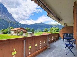 A balcony with a view of mountains, a wooden fence with decorative elements, and a table with blue chairs. - Firstsunne by Interhome (Grindelwald)