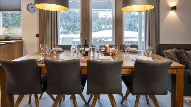 Un coin repas avec une table en bois dressée pour quatre personnes, entourée de chaises, et de grandes fenêtres donnant sur un paysage enneigé.