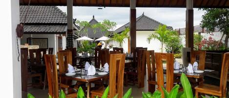 Un área de comedor al aire libre con mesas y sillas de madera, sombrillas y plantas tropicales.
