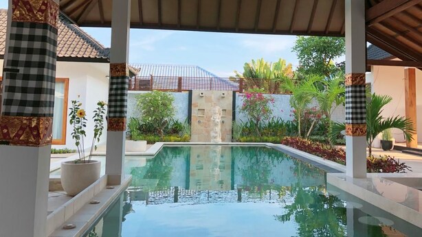 A pool area with a clear view of the pool, surrounded by white pillars with patterned bases, a tiled roof, and a stone wall with a gate. - Villa Casablanca Surf Keramas (Gianyar)