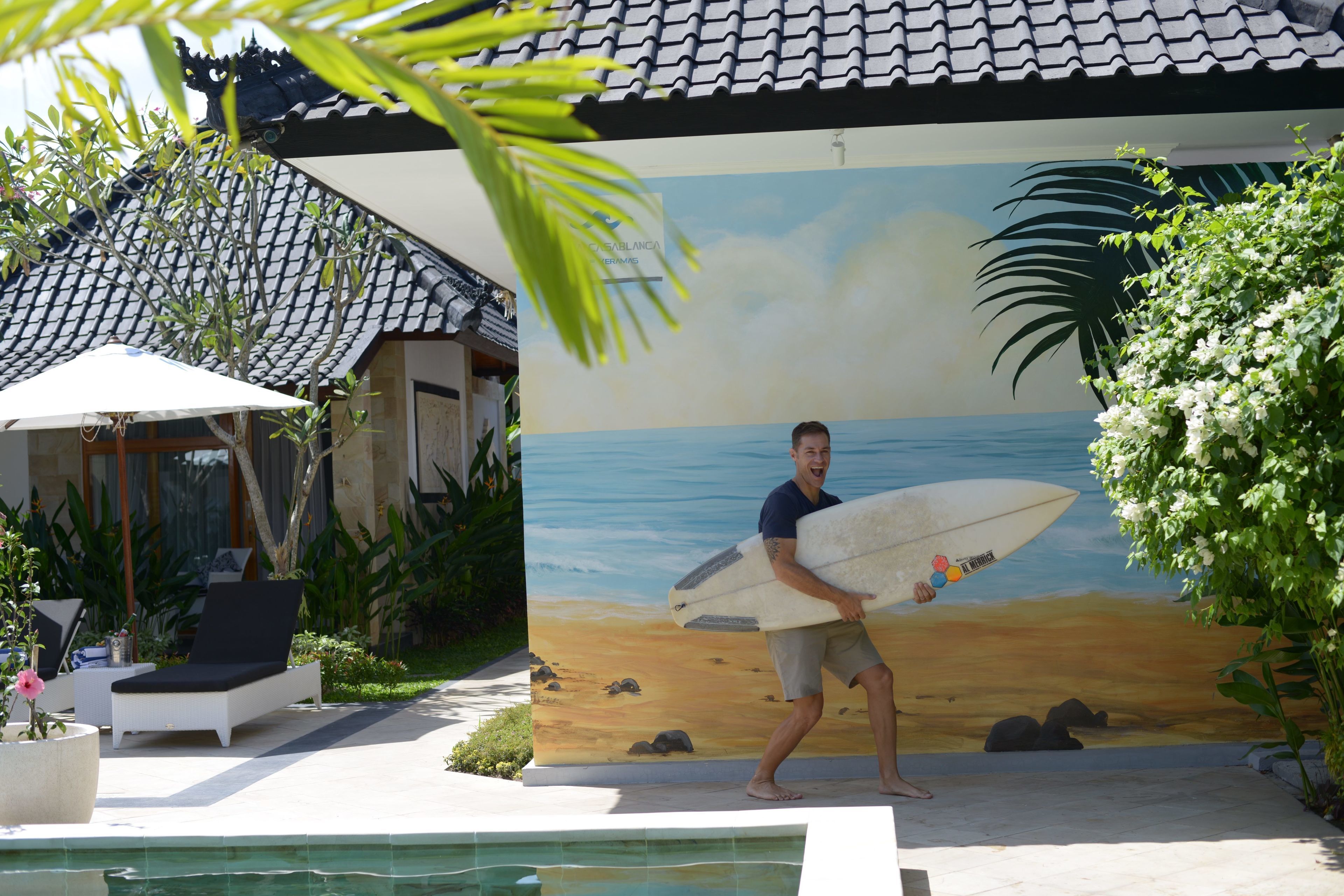 A man carrying a surfboard, a pool, and a mural of a beach scene.
