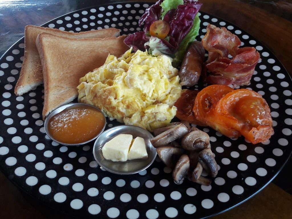 A breakfast plate with toast, scrambled eggs, bacon, mushrooms, and a side of butter and jam.