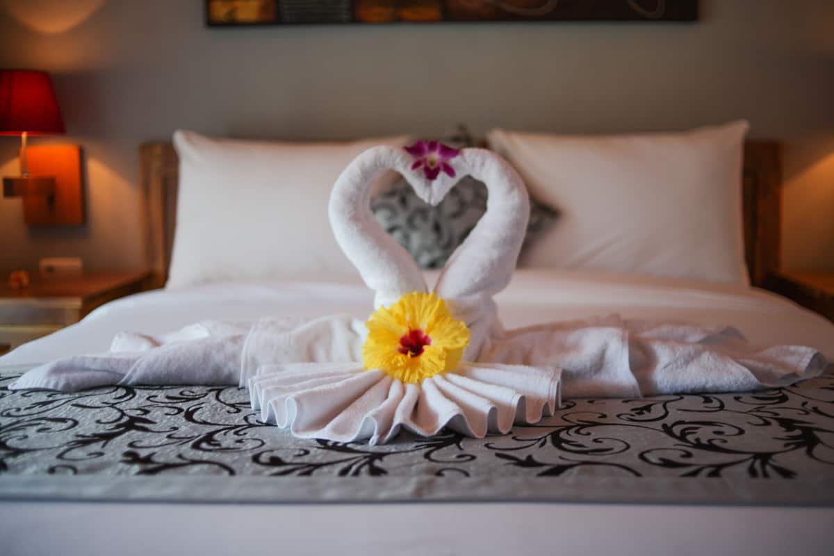 A neatly made bed with a heart-shaped towel arrangement and a yellow flower centerpiece.