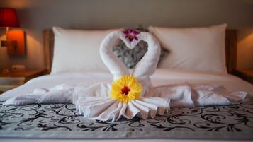Una cama cuidadosamente hecha con una toalla dispuesta en forma de corazón y un centro de mesa con una flor amarilla.
