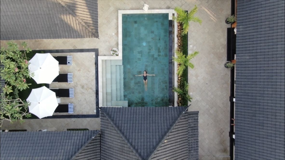 Vista aérea de uma piscina com uma pessoa na água, rodeada por guarda-sóis e plantas em vasos.
