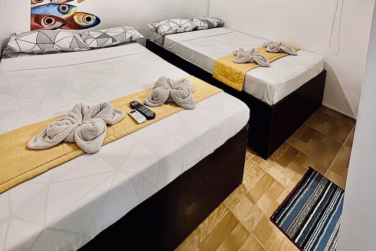 A hotel room with two beds, a ceiling fan, a painting of fish, a window with blinds, and a small rug.