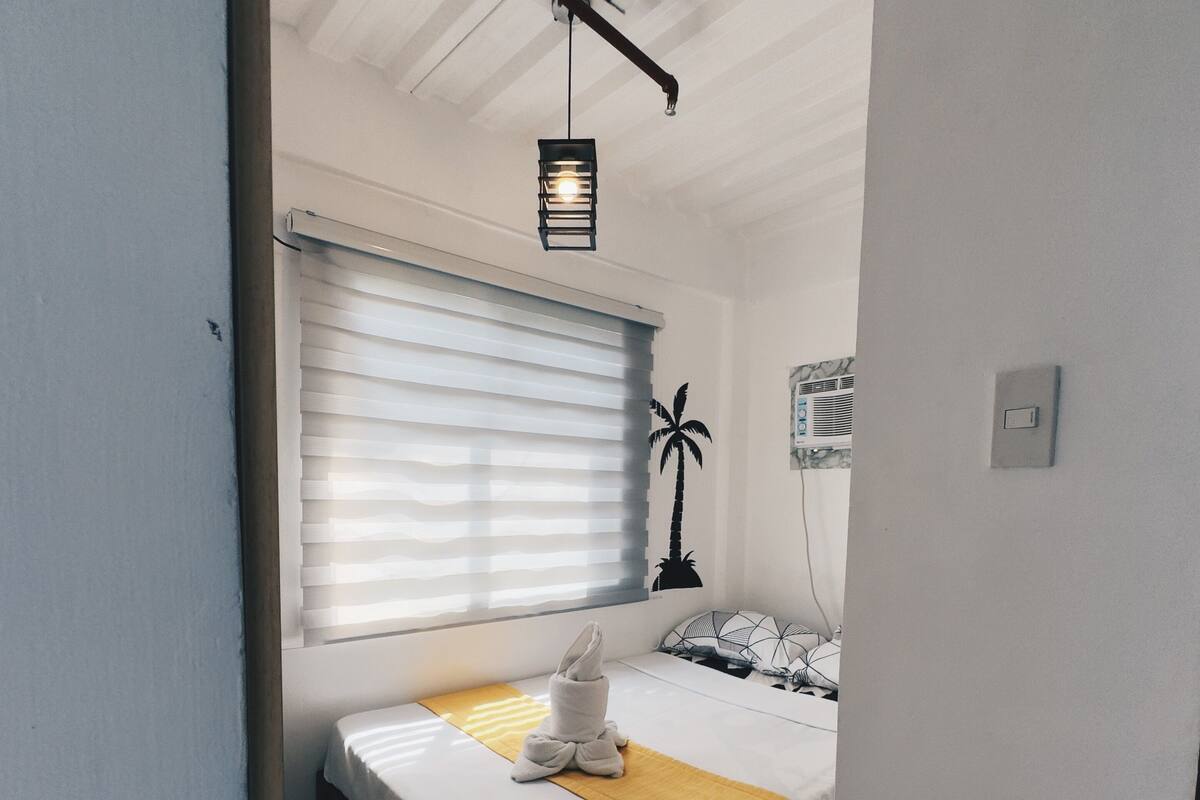A compact bedroom with a bed, a bedside table, a lamp, and a window with blinds.