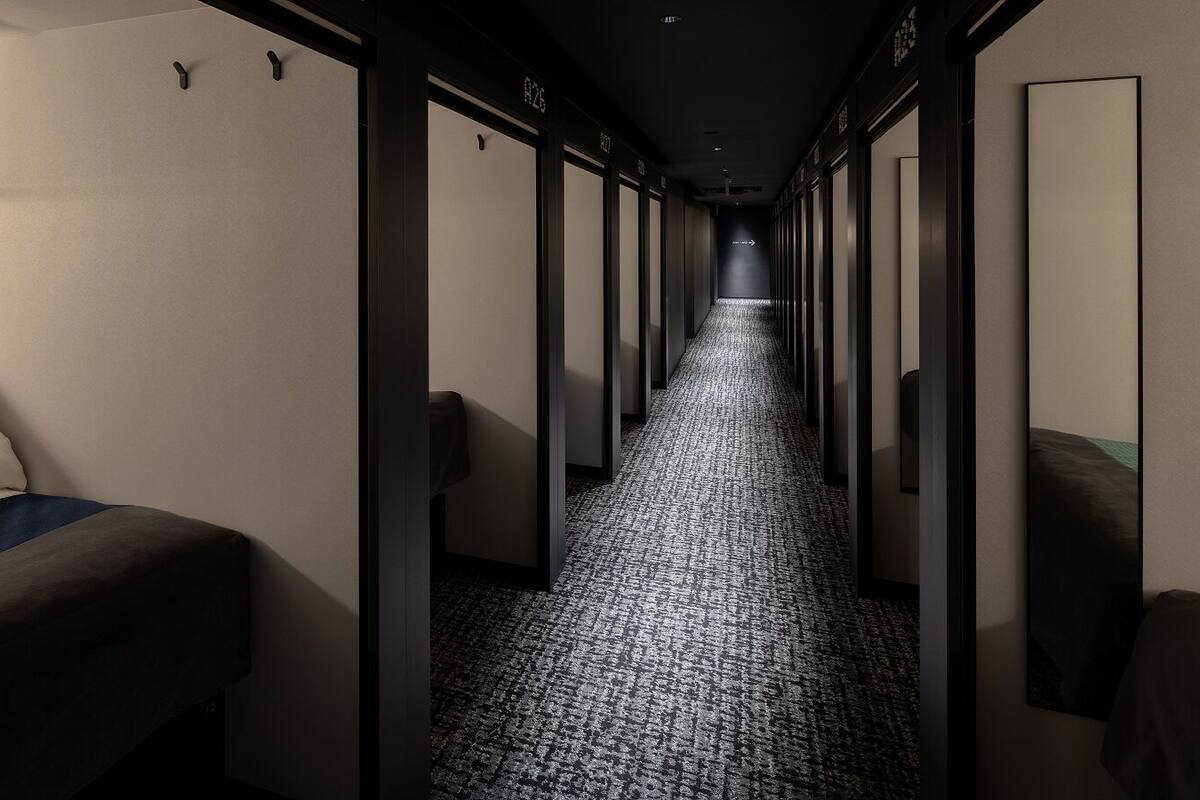 Un couloir d’hôtel avec un sol recouvert de moquette, un lit avec une couvre-lit bleue et une rangée de portes sur le côté droit.