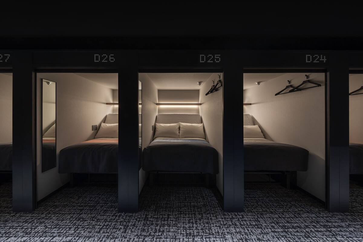 Une chambre d’hôtel avec trois lits, chacun étant identifié par un numéro (D26, D25, D24). La chambre est dotée d’un sol recouvert de moquette et d’un miroir au mur.