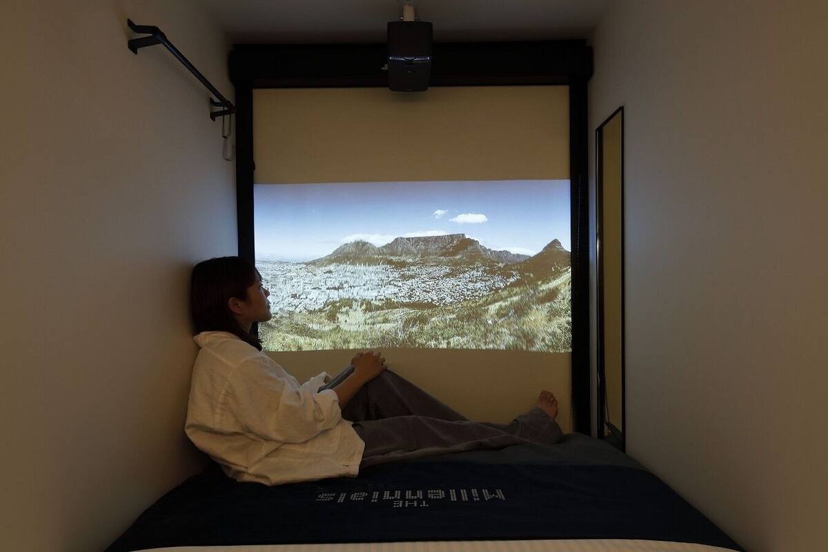 Une personne assise sur un lit, dans une pièce où un écran de projection diffuse un paysage.