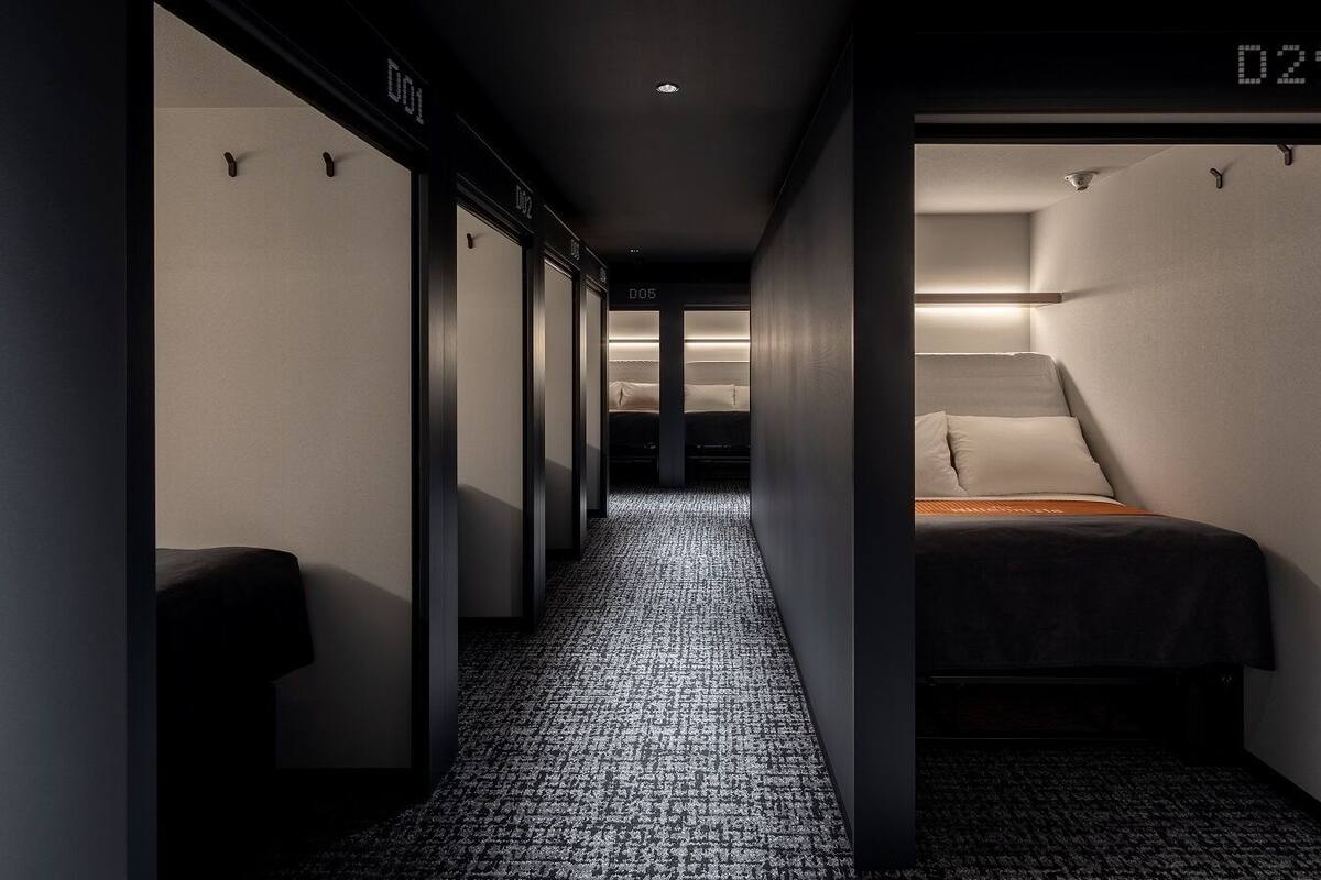 Un couloir étroit avec un tapis à motifs, un lit avec un matelas et des oreillers, et une horloge numérique affichant l’heure.