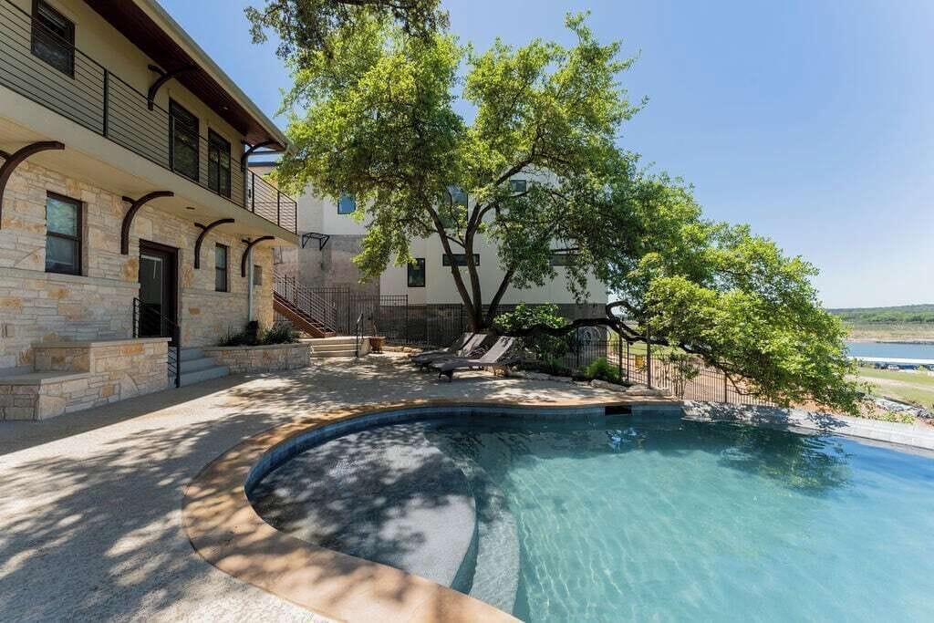 Una piscina con borde curvo, un edificio de piedra con balcón y un árbol que da sombra.