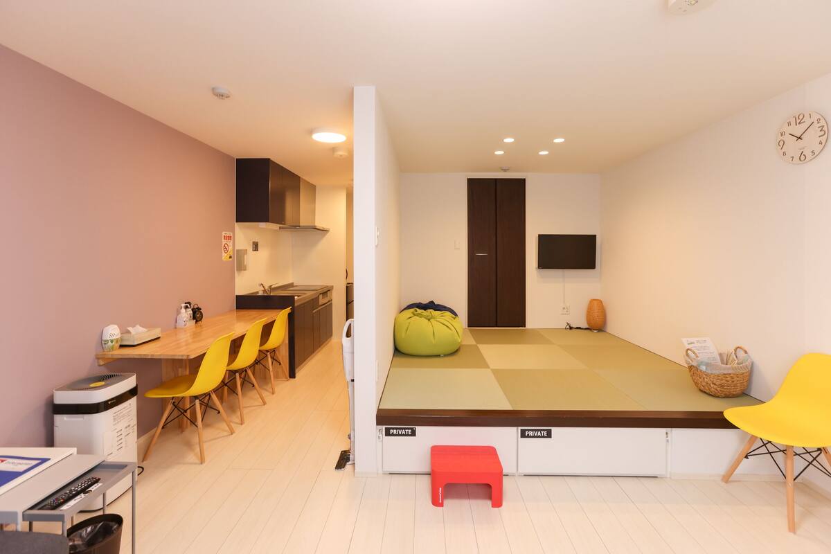Een compacte kamer met kitchenette, een bed met opbergruimte, een televisie en een klein zithoekje met stoelen.