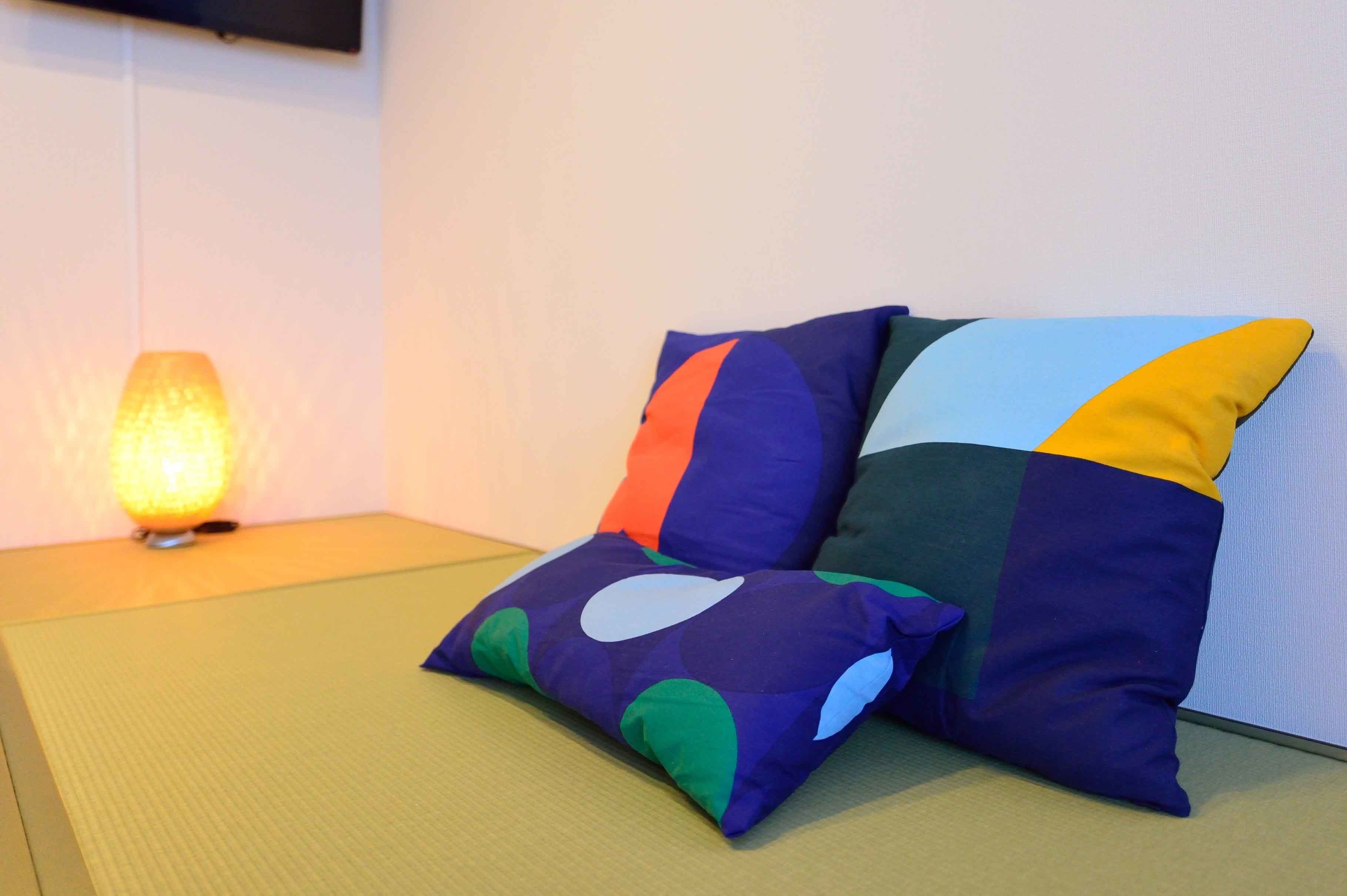 A room with tatami flooring, a wall-mounted TV, and two colorful pillows.