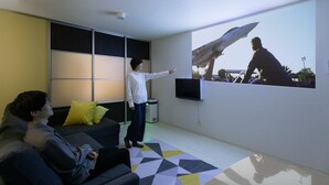 Una sala de estar moderna con una pantalla grande mostrando una imagen de un avión de combate, un sofá, una mesa de centro y una televisión.
