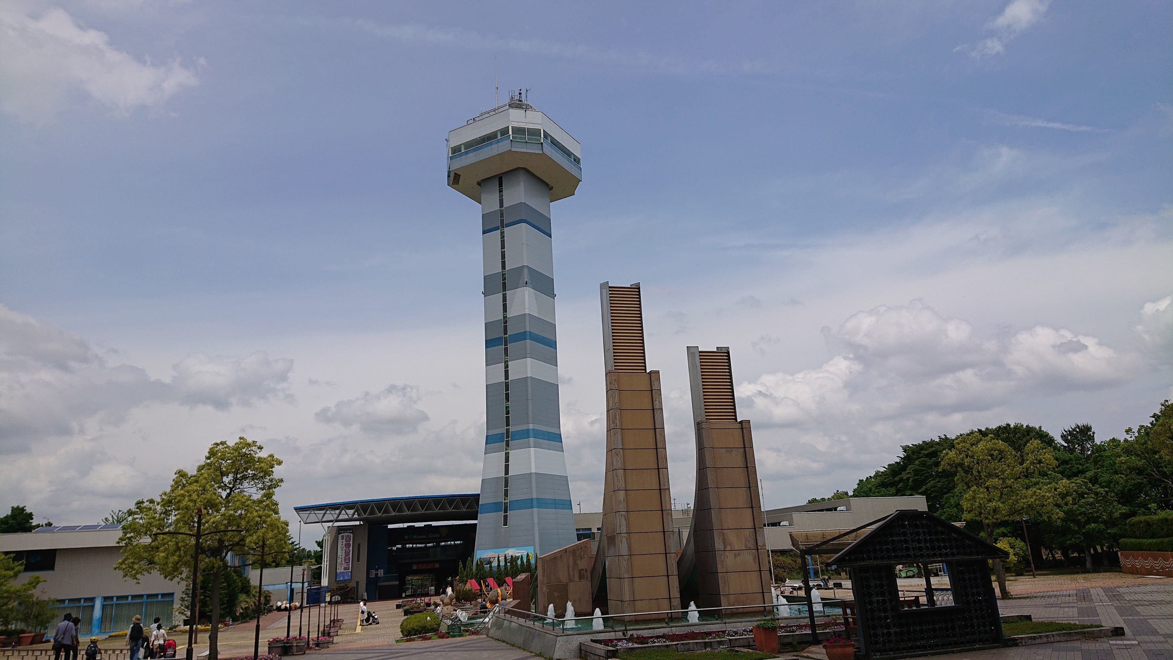 A tall tower with a control room at the top, a large open plaza with a fountain, and a building with a blue and white facade.