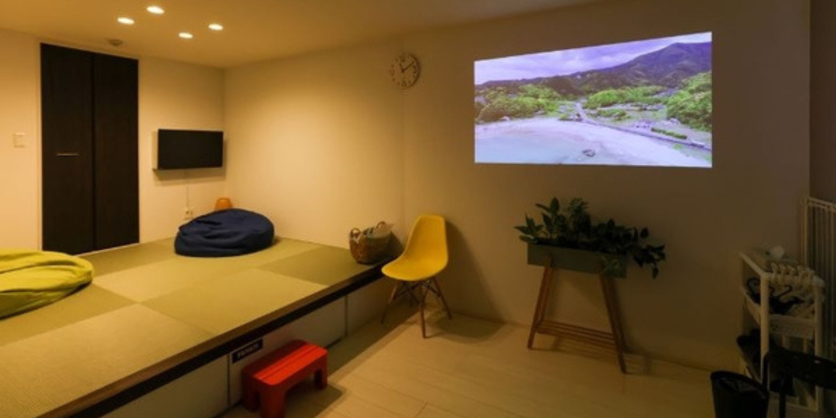 Una habitación con cama, televisión, pantalla de proyección, una silla amarilla y una planta pequeña.