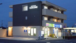 Un edificio moderno con un letrero que dice "GUESTHOUSE COCONE". Cuenta con un letrero de estacionamiento, una cabina de venta de boletos y un letrero para una cafetería.