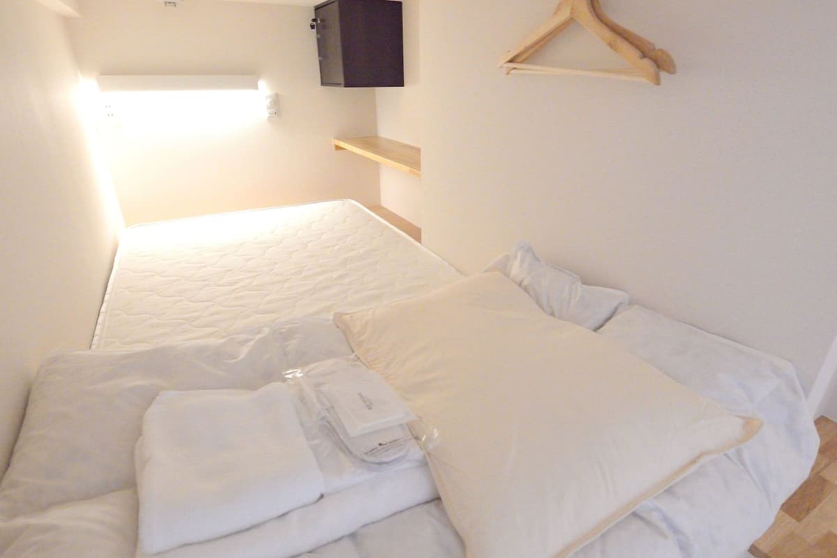 Een netjes opgemaakt bed met kussens en een opgevouwen handdoek, een houten kleerhanger en een klein plankje met een flatscreen televisie aan de muur.