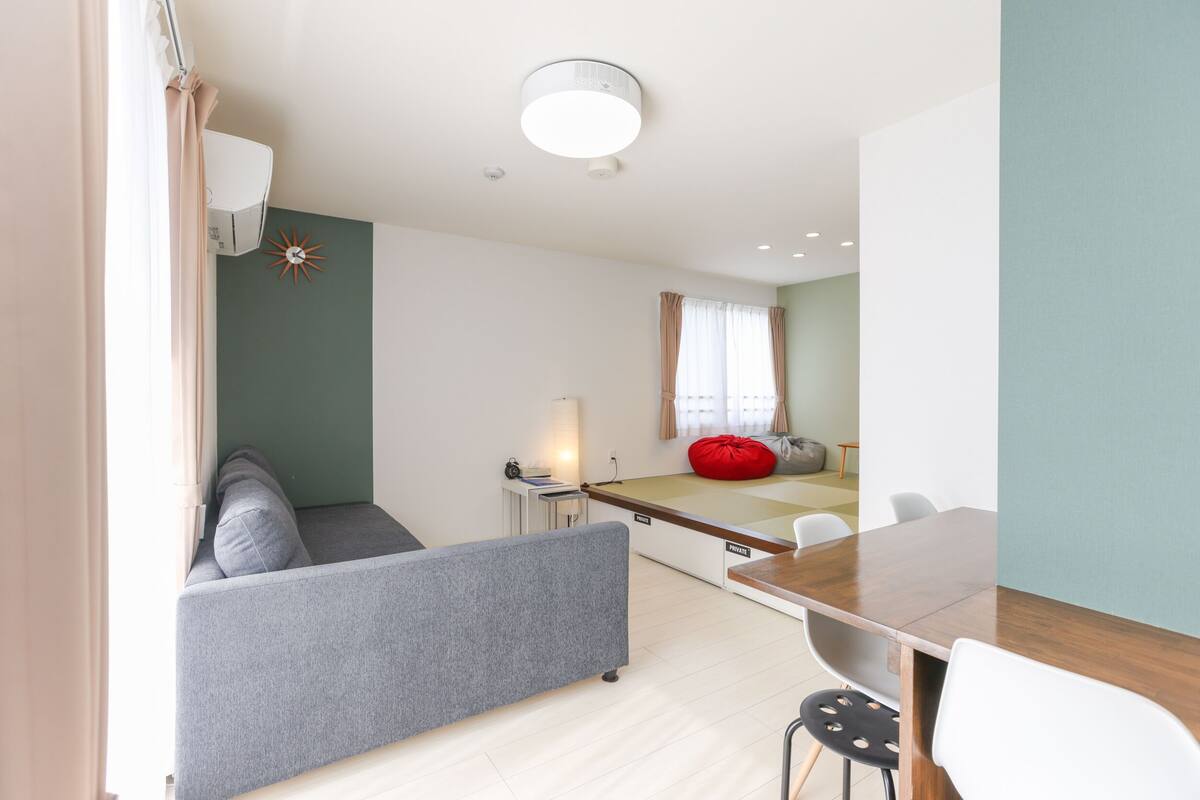 Een moderne woonkamer met een grijze bank, een eettafel met stoelen, een rode relaxstoel en een klein bijzettafeltje met een lamp.