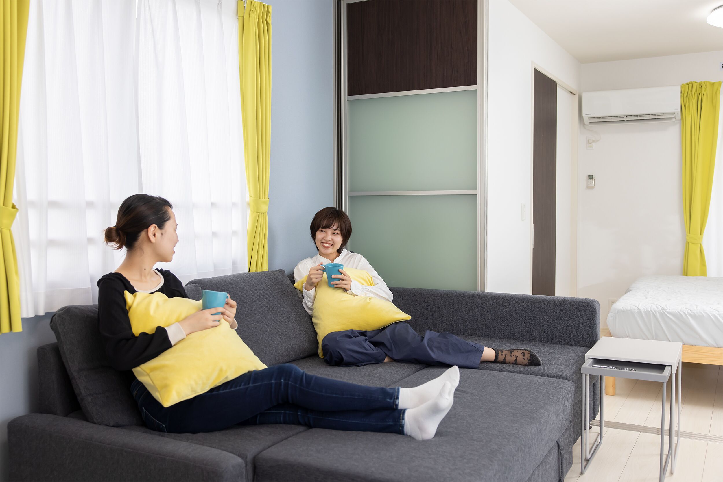 A modern living room with a grey sofa, yellow cushions, a small table, and a bed in the background.