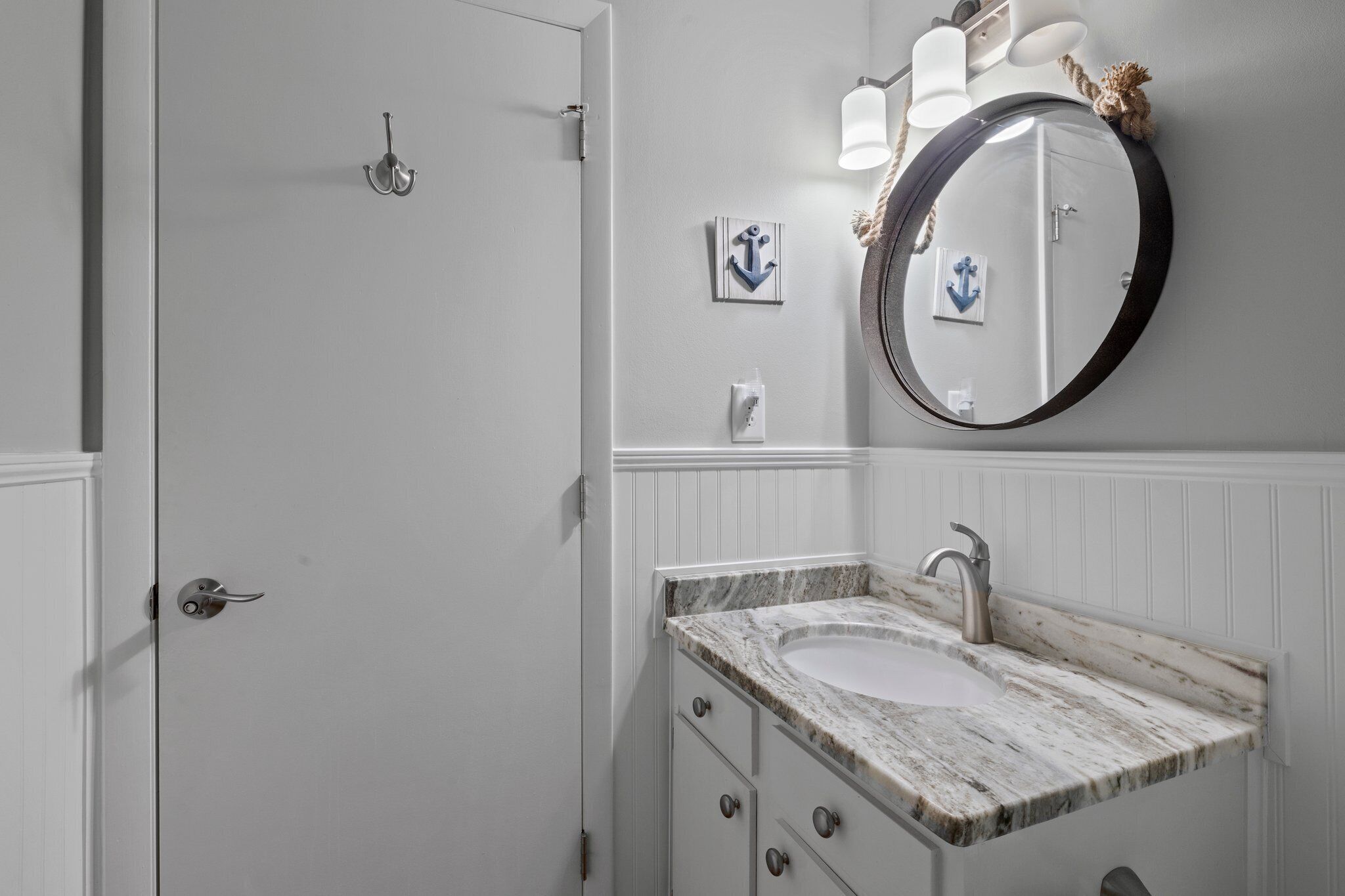 A bathroom with a white door, a marble sink, a round mirror with a rope decoration, and an anchor artwork on the wall.
