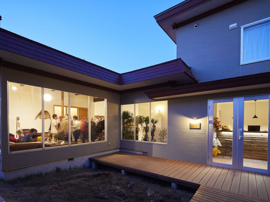 Een modern huis met grote glasramen, een houten terras en een zichtbare keukenruimte.