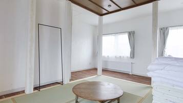 Quarto com piso de madeira, uma mesa redonda, cama com roupa de cama branca e uma janela com cortinas.