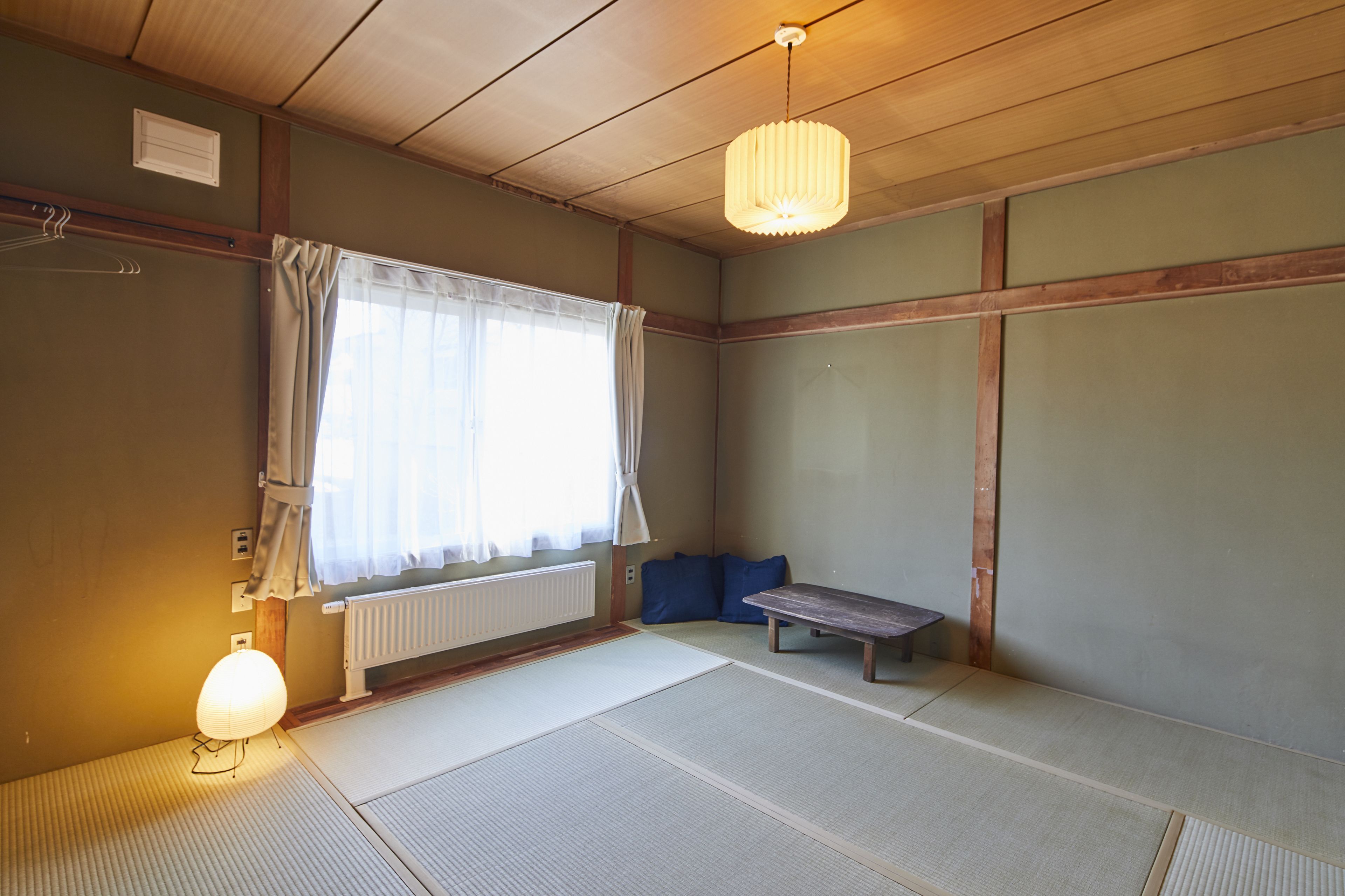 A traditional Japanese room with tatami flooring, a low wooden table, a hanging light fixture, and a window with sheer curtains.