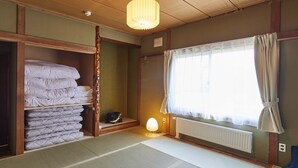 A traditional Japanese room with tatami flooring, a wooden wardrobe, a stack of futons, a radiator, and a window with curtains. - haku hostel (Shiraoi)