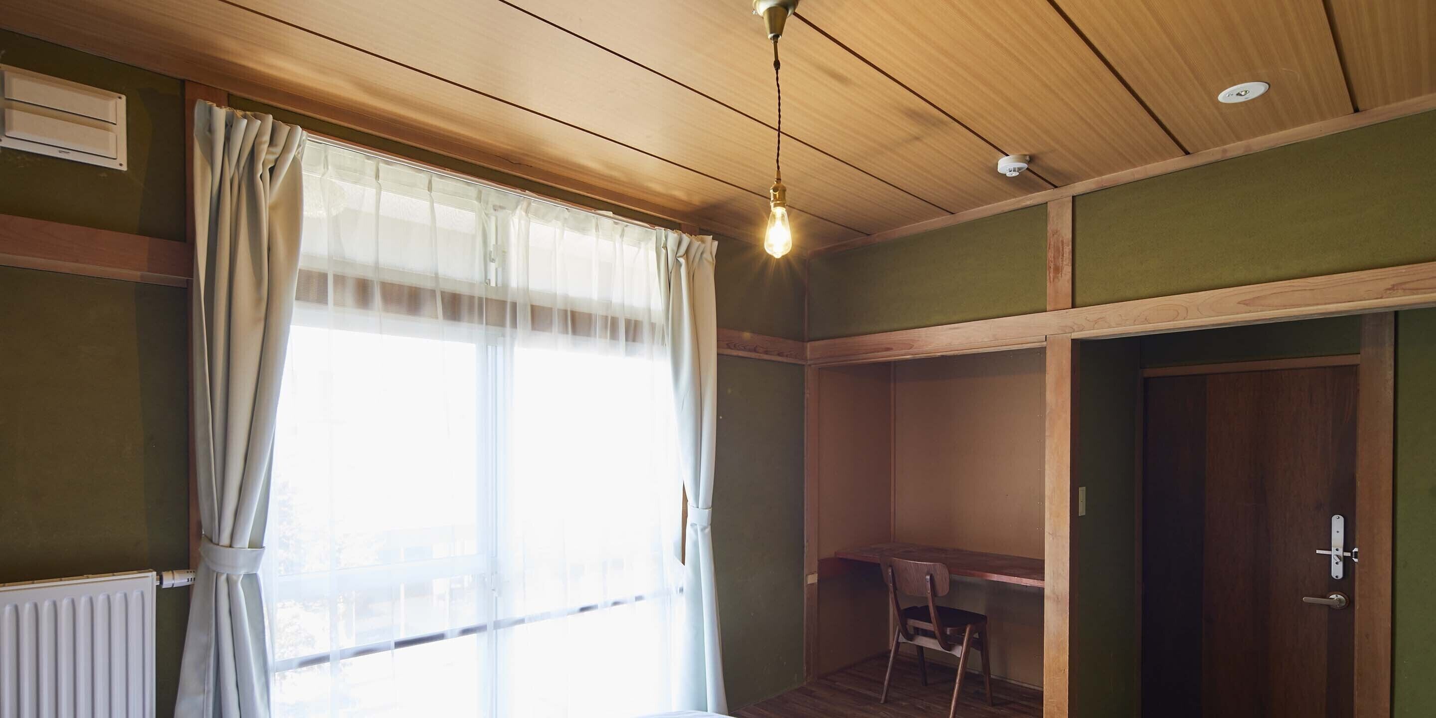 A room with a wooden ceiling, a radiator, a window with curtains, a desk, and a chair.