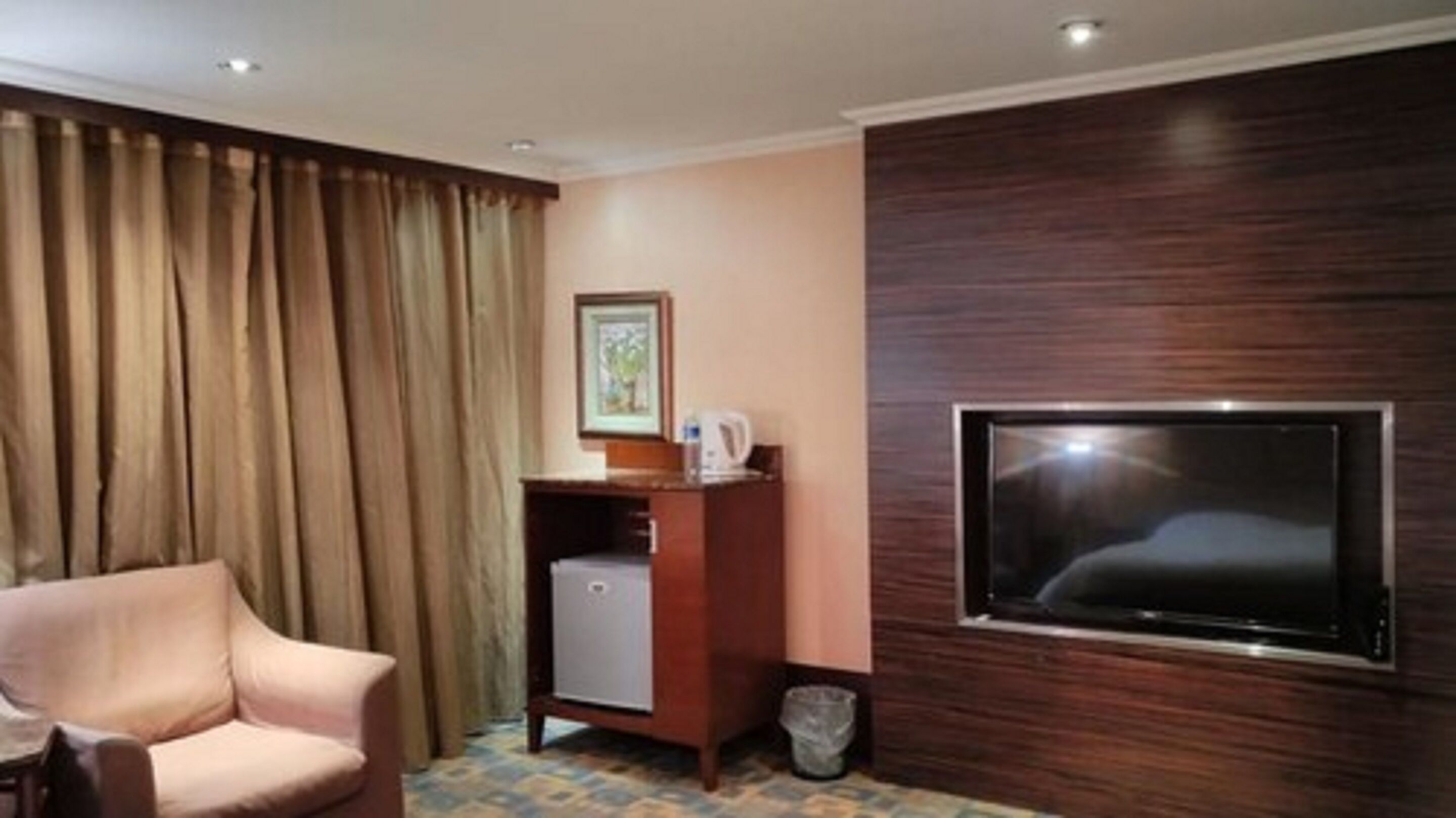 A hotel room with a flat-screen TV mounted on a wooden wall, a small wooden cabinet with a picture frame and a kettle, a beige armchair, and a curtain.