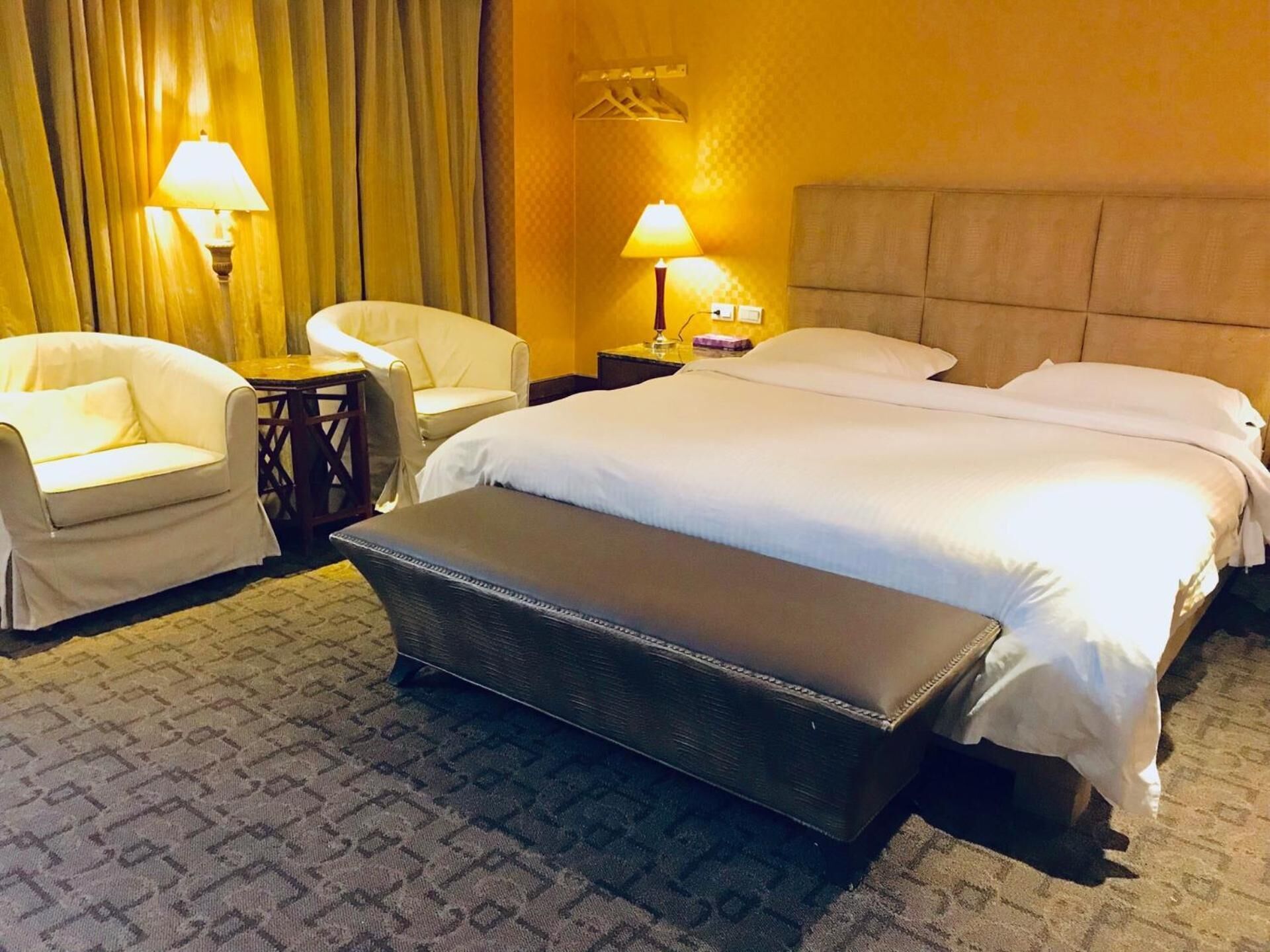 A hotel room with a large bed, two bedside tables, two lamps, a chair, and a bench.