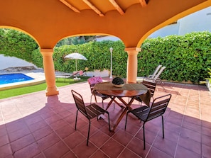 Un patio cubierto con piscina, mobiliario exterior y un seto verde y frondoso.
