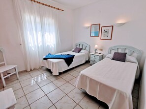 Habitación de hotel con dos camas, una silla, una mesita de noche con lámpara y una ventana con cortinas translúcidas.