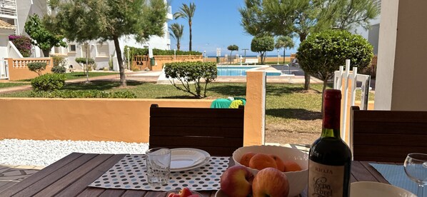 Una mesa con una botella de vino Rioja, un bol de naranjas y un plato con fruta, con vistas a una piscina y un área ajardinada con árboles y vista al mar.