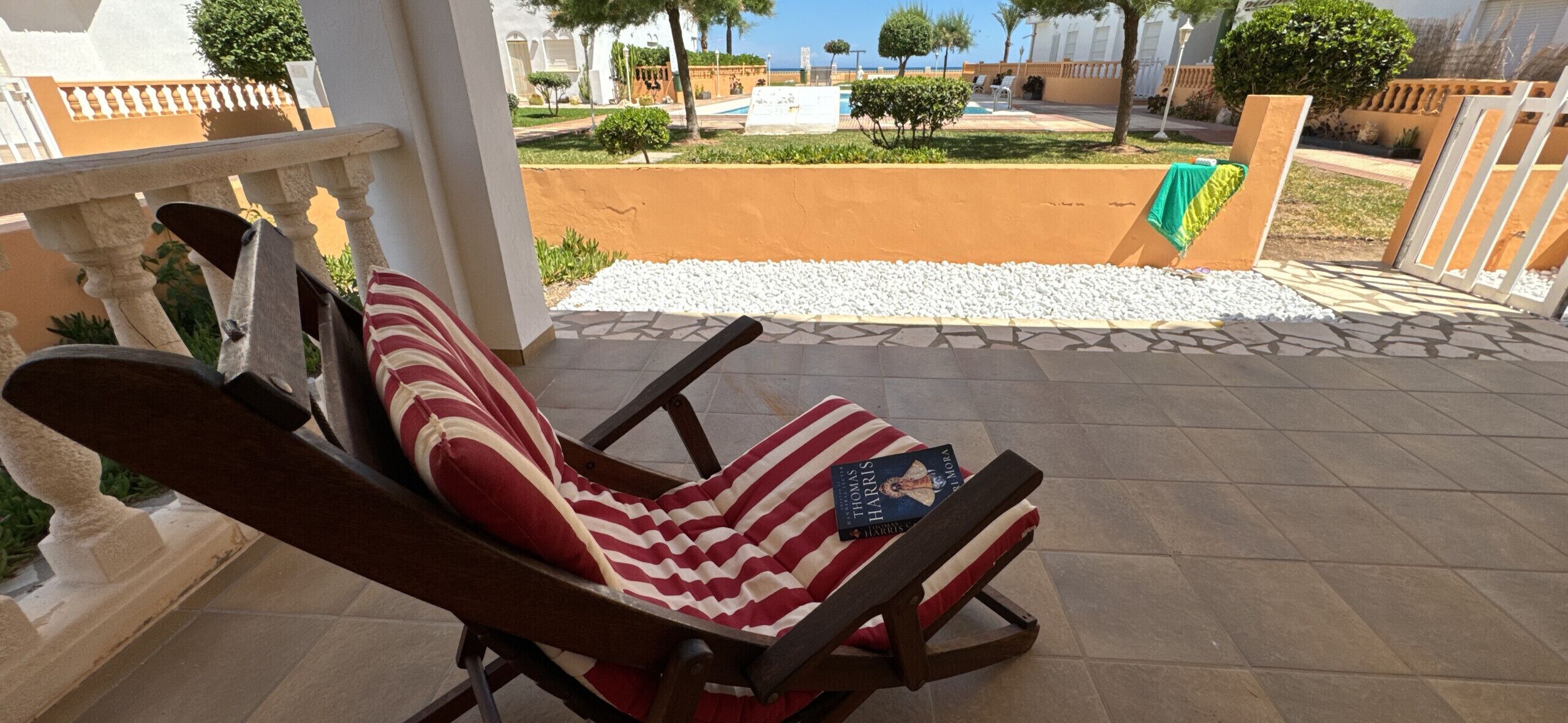 A wooden lounge chair with a striped cushion and a book on it, positioned on a tiled patio with a view of a landscaped garden and a pool.