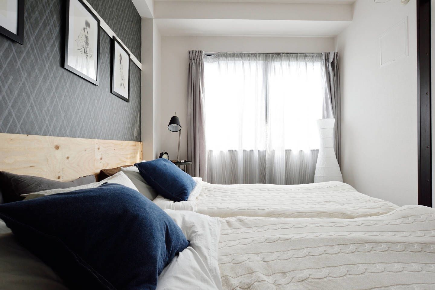 A double bed with white bedding and blue pillows, a wooden headboard, a bedside table with a lamp, and a window with sheer curtains.