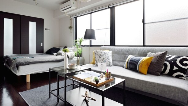 A modern, well-lit bedroom with a large window, a bed, a sofa, and a small table with a lamp and decorative items. - nid NIPPONBASHI at Ebisu (Osaka)