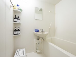 Ein kleines, weißes Badezimmer mit Waschbecken, Spiegel und Dusche. Es gibt Ablagen mit Pflegeprodukten und eine Treppenstufe mit Punkten.