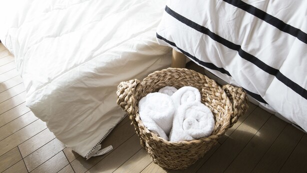 Una cama bien hecha con ropa de cama blanca y una almohada a rayas, una cesta de mimbre con toallas dobladas y un piso de madera.