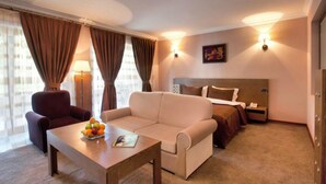 Quarto de hotel com sofá, poltrona, mesa de cabeceira, luminária e cama com encosto.